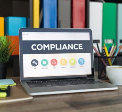 Laptop on a desk displaying the word 'Compliance' on the screen. Surrounding the laptop are a cup of coffee, a small potted plant, colorful sticky notes, and a pencil holder.