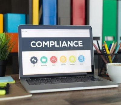 Laptop on a desk displaying the word 'Compliance' on the screen. Surrounding the laptop are a cup of coffee, a small potted plant, colorful sticky notes, and a pencil holder.